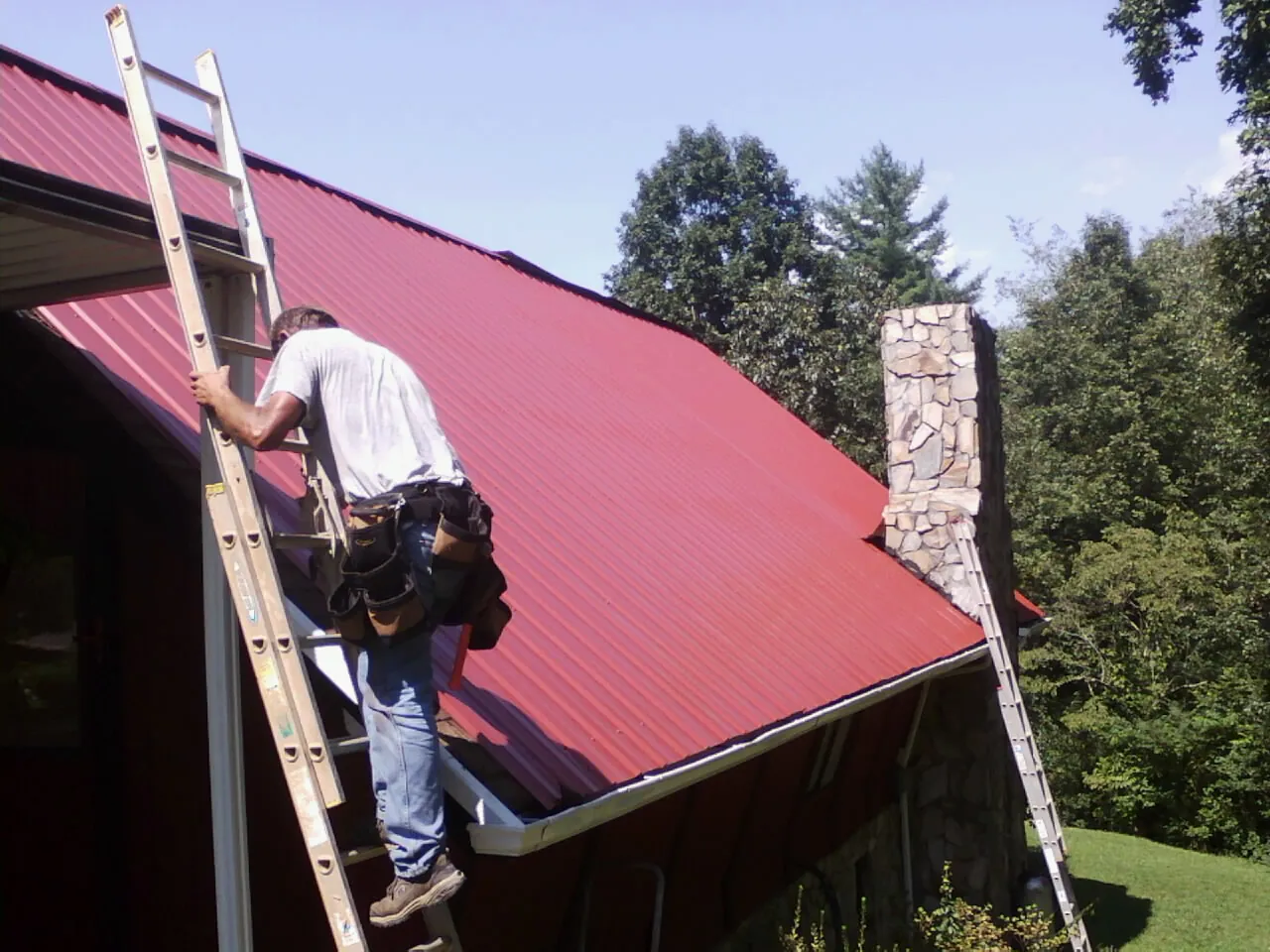 Roofer on a ladder beside a red metal roof and stone chimney — Soffit & Fascia Repair by Roofing Brothers in Denton